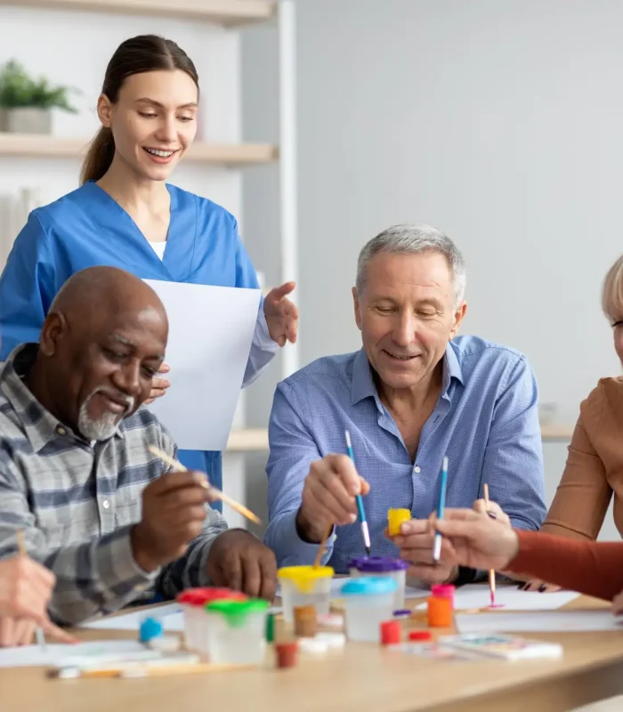 Happy multiracial elderly men and women sitting around table and drawing at nursing home, doing arts and crafts together, young lady nurse assisting group of senior people, copy space