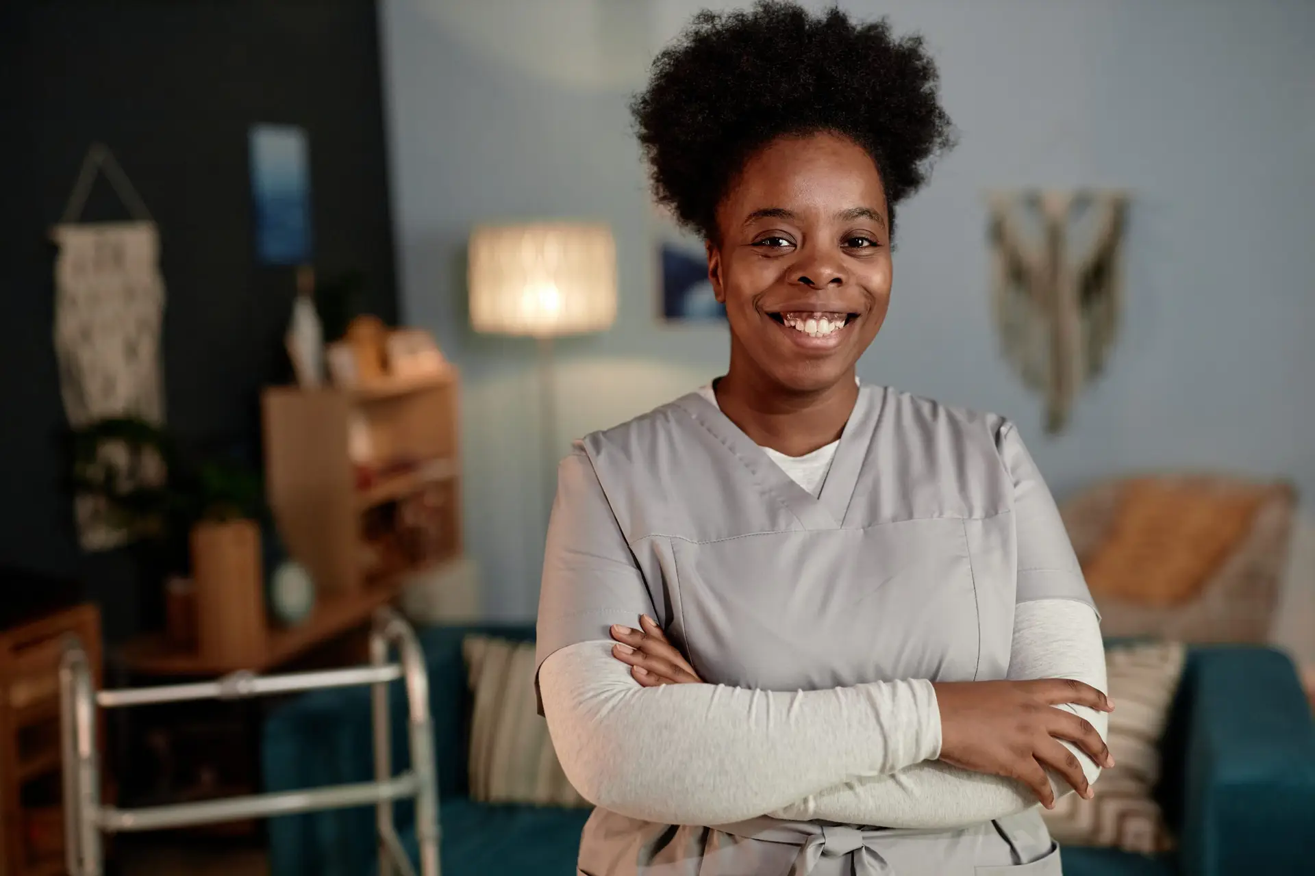 Medium portrait of African American female social worker beaming with smile wearing scrubs posing with arms crossed during house visit to patient, copy space